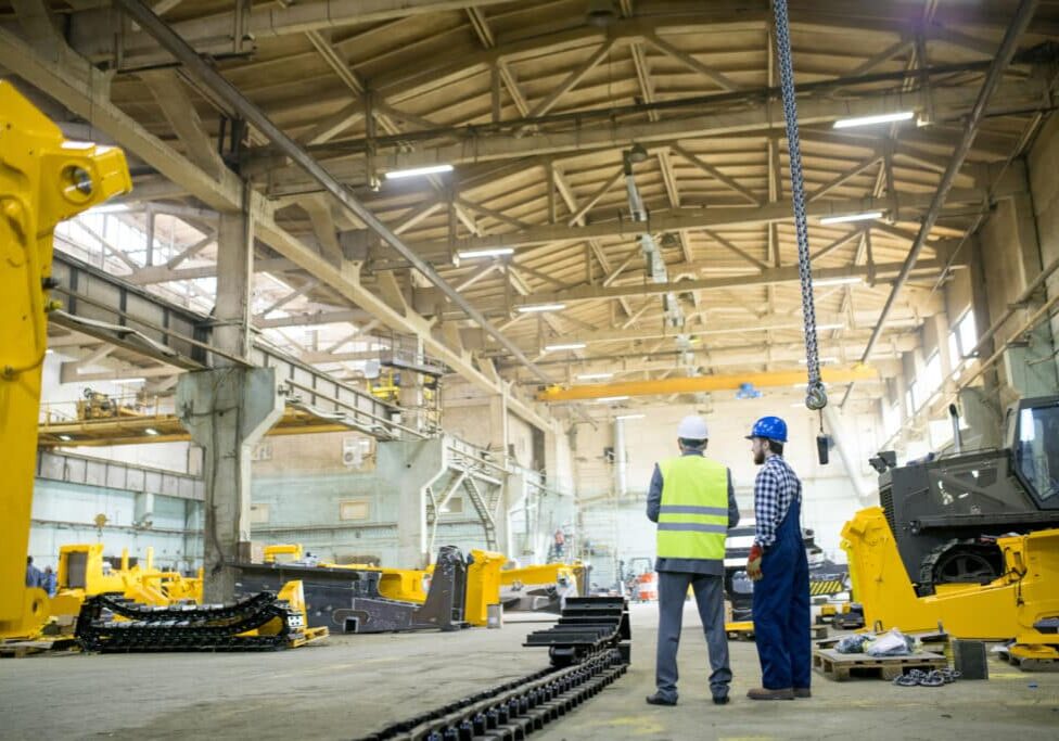 Rear view of production coordinator working with main engineer at assembling workshop of factory, they discussing tractor parts Production coordinator working with main engineer